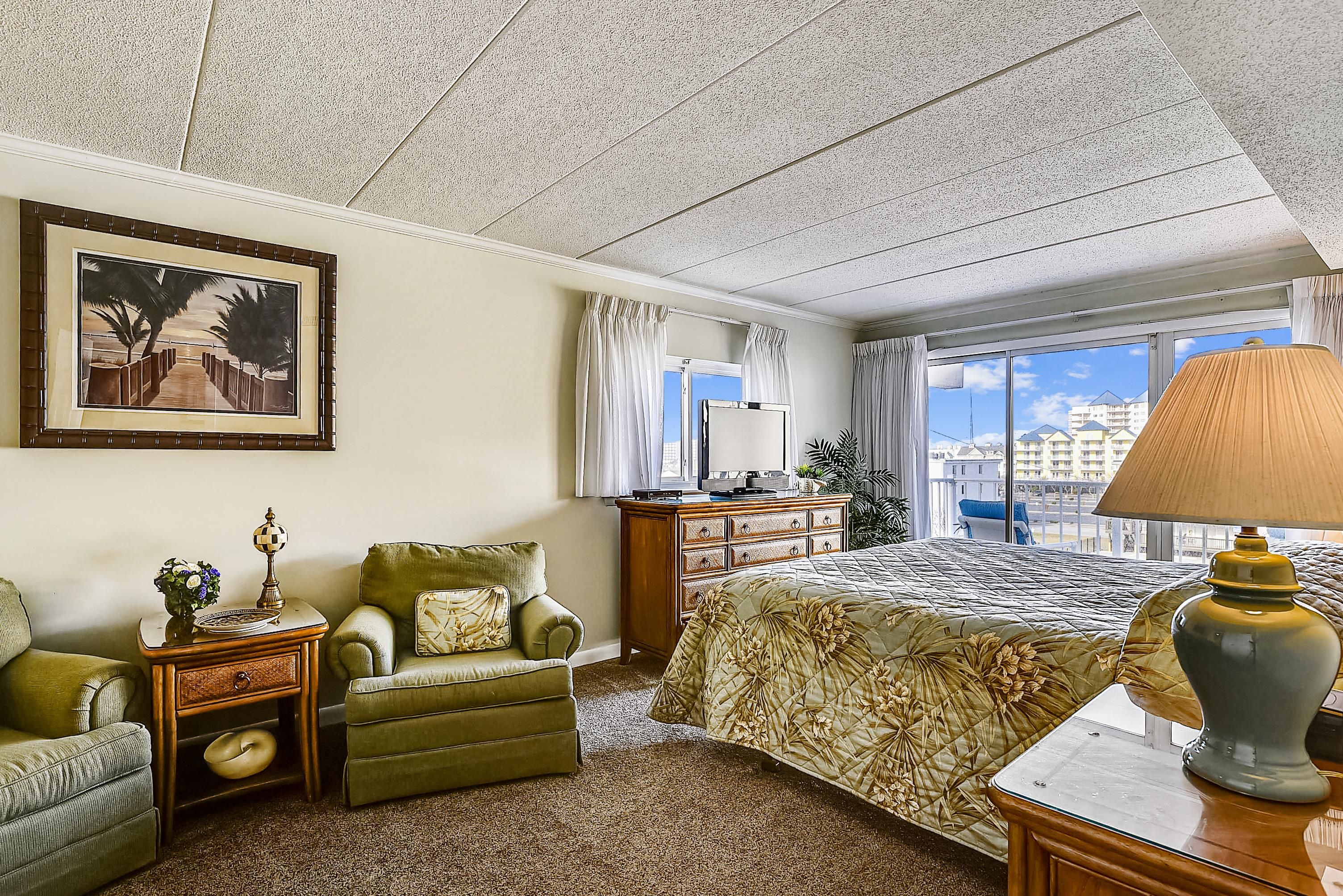 A hotel room with a large bed, a desk, a chair, a dresser, a painting, and a view of the city.