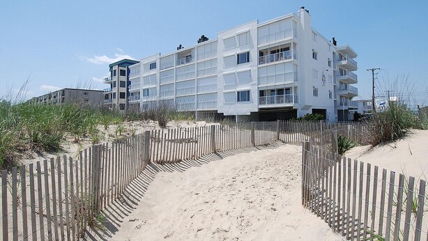 A multi-story building with a sandy pathway leading to it, surrounded by a wooden fence.