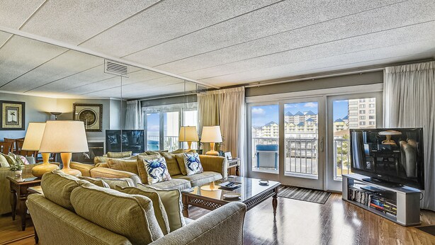 A spacious living room with a sofa set, a coffee table, a television, and a large window with a city view. - DAILY ACTIVITIES & LINENS INCLUDED! 3BR, 2BA Condo with a Large Balcony. Great mid-town location! (Ocean City)