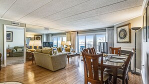 A spacious living room with a large sofa, dining table, and chairs, a television, and a view through large windows. - DAILY ACTIVITIES & LINENS INCLUDED! 3BR, 2BA Condo with a Large Balcony. Great mid-town location! (Ocean City)