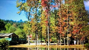 Una cabaña junto a un lago con un reflejo nítido, rodeada de árboles con follaje otoñal.
