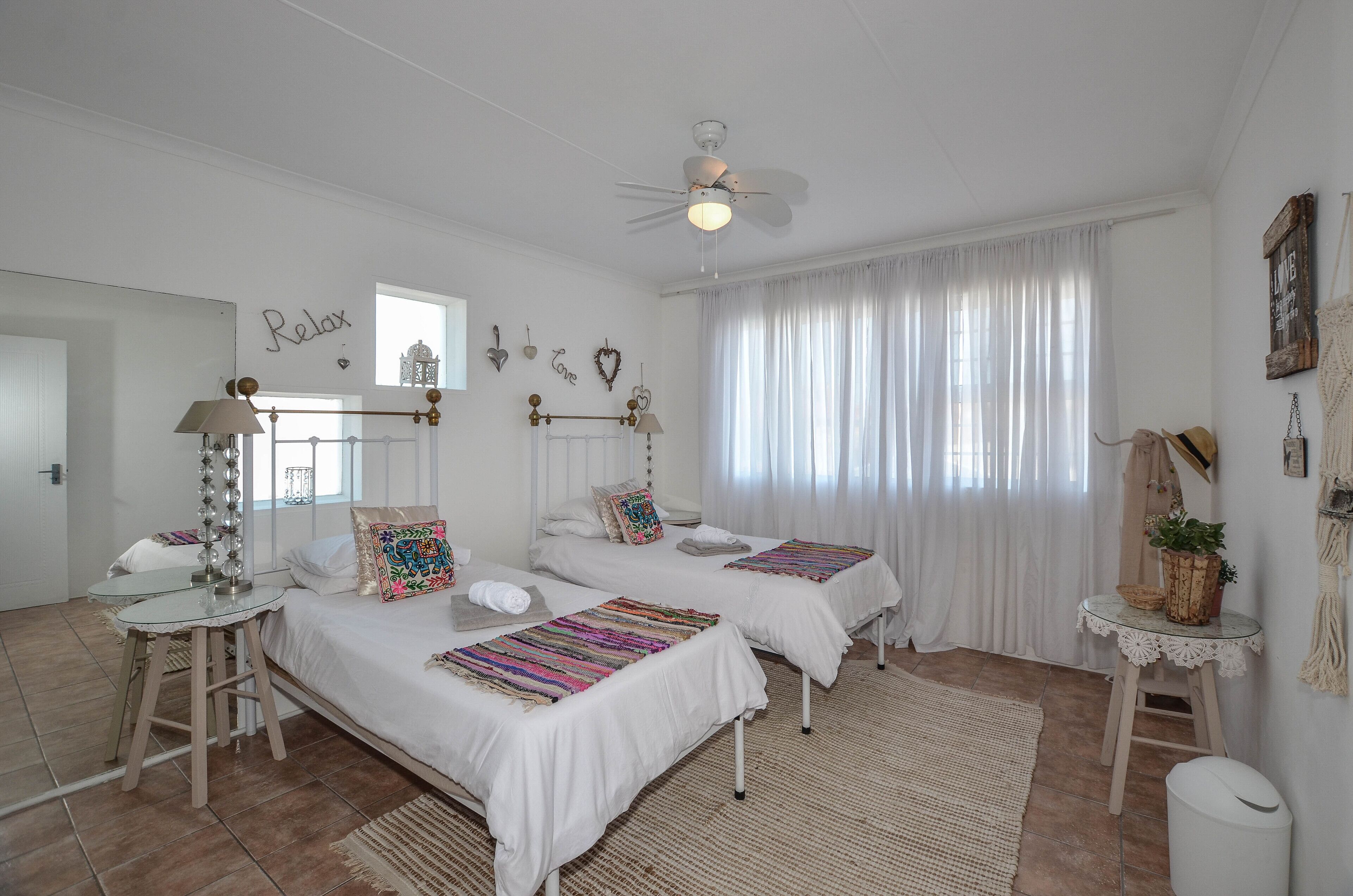 Un dormitorio bien iluminado con dos camas, un ventilador de techo, una mesita pequeña y una ventana con cortinas.