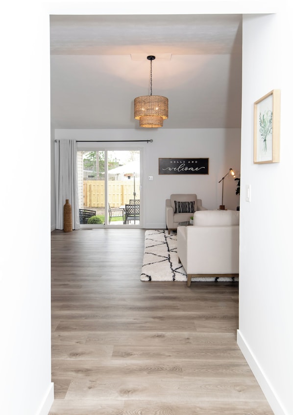 A modern living room with a wooden floor, a white sofa, a wicker pendant light, and a sliding glass door leading to a backyard.