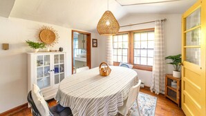 Un coin repas comprenant une table ronde, des chaises, un luminaire suspendu, un meuble avec des portes en verre et une fenêtre avec des rideaux.