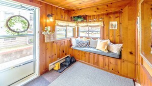 L’intérieur d’un chalet accueillant, avec un décor en bois, un banc avec des coussins, une fenêtre ornée d’une guirlande et une porte.