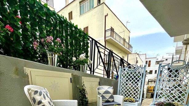 Un’area patio con mobili in vimini, tra cui un divano e un tavolino rotondo, circondata da piante in vaso e un recinto decorativo.