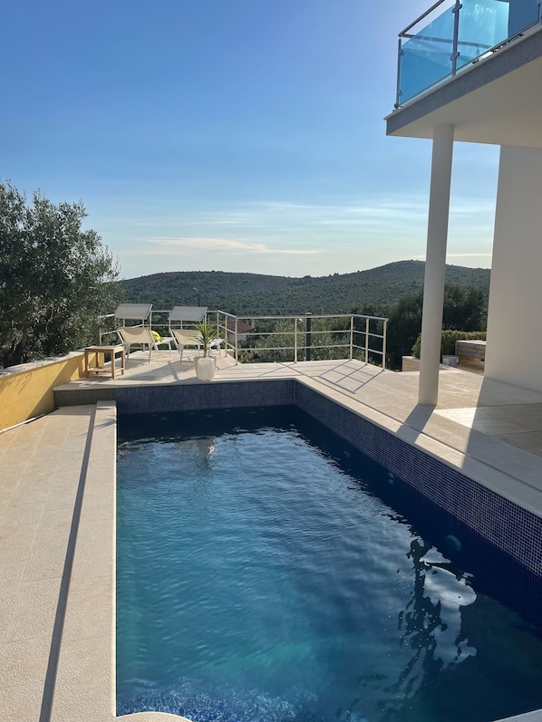 A swimming pool with a view of a hilly landscape, surrounded by a modern outdoor area with lounge chairs and a glass balustrade.