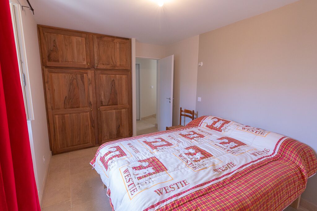 A bedroom with a large wooden wardrobe, a bed with a quilt featuring the name 'WESTIE', and a red curtain on the left.