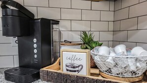 Een aanrecht met een koffiezetapparaat, een bord met een koffie-spreuk, een mand met witte bekers en een kleine potplant.