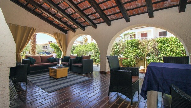 An outdoor patio with a brick floor, covered by a wooden ceiling, furnished with sofas and chairs, and surrounded by arches.