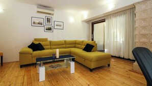 A living room with a yellow sofa, a glass coffee table, and a black armchair. There are framed pictures on the wall and a window with curtains.