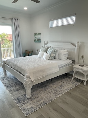 A bedroom with a white bed, a nightstand, a small table, a ceiling fan, a window with blinds, and a painting on the wall.