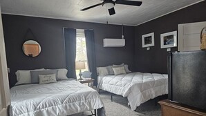 A room with two beds, a ceiling fan, an air conditioning unit, a refrigerator, and a small table with a lamp.