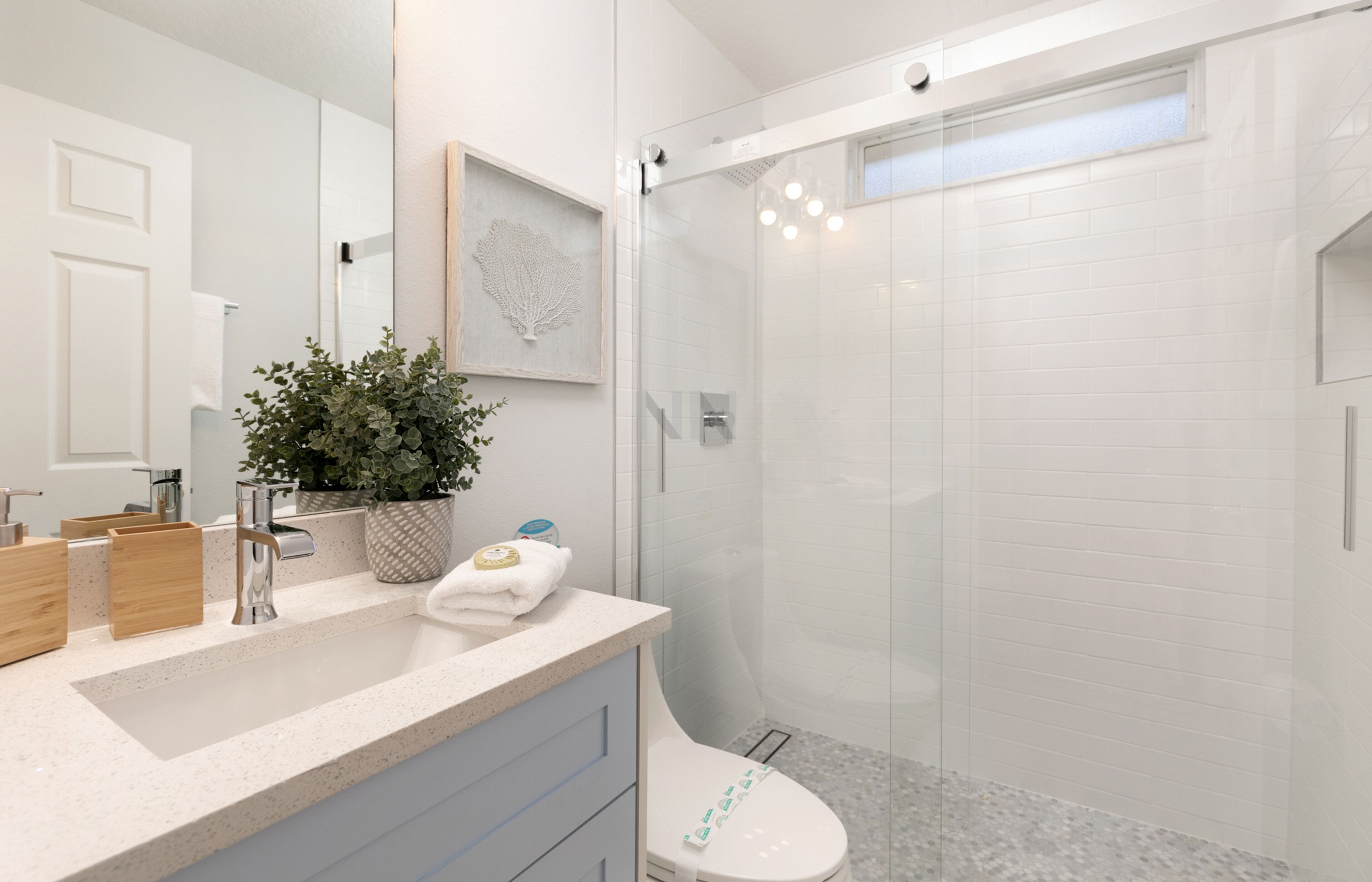Un baño moderno con ducha de vidrio, un lavamanos blanco con una planta y un espejo grande.