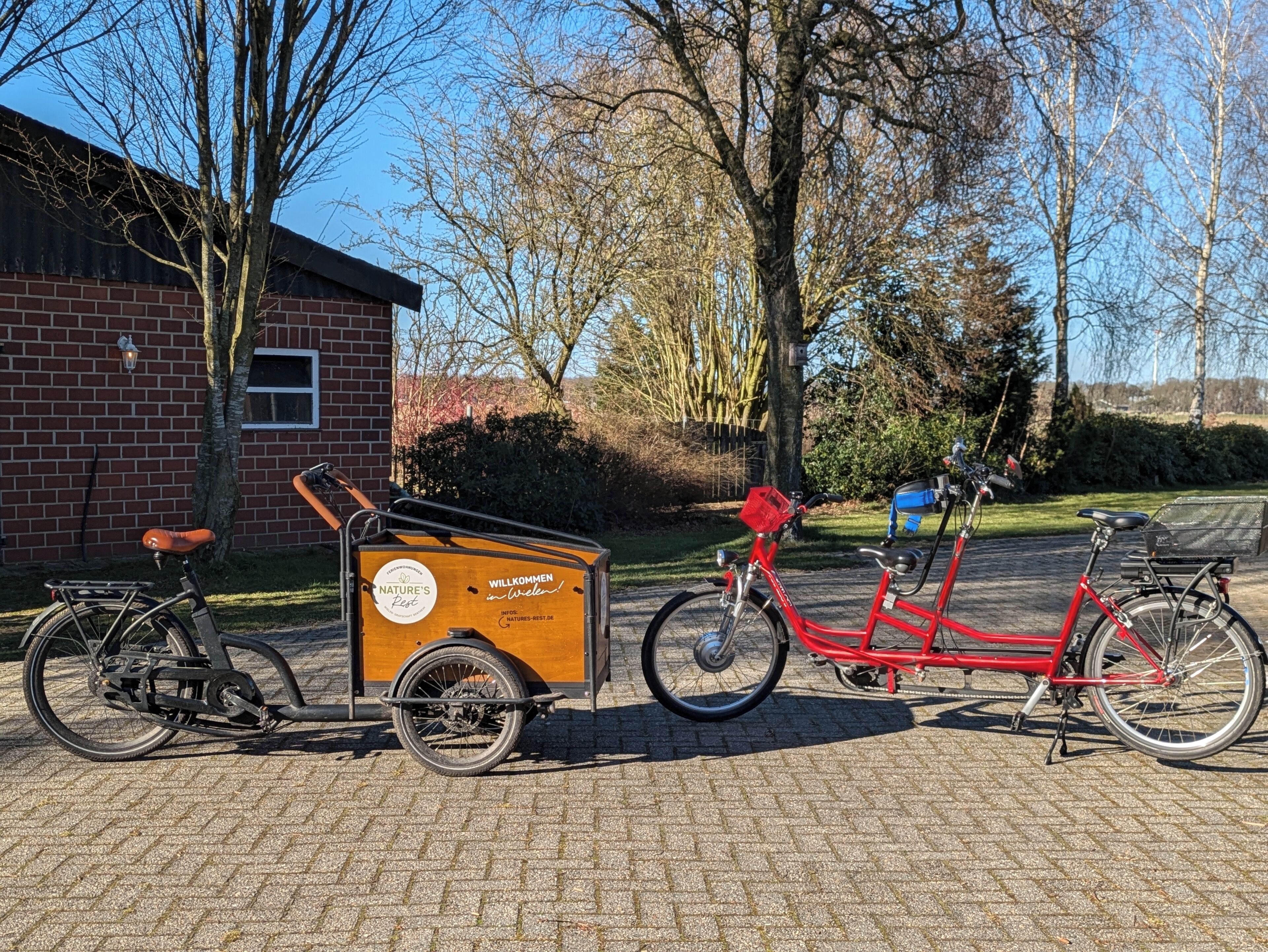 Two bicycles parked on a paved area with a brick building and trees in the background.