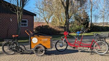 Two bicycles parked on a paved area with a brick building and trees in the background.