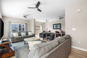 Un salón amplio con un sofá, sillones, una mesa de centro y un televisor. Hay ventiladores de techo, un comedor con sillas y un gran ventanal con cortinas.