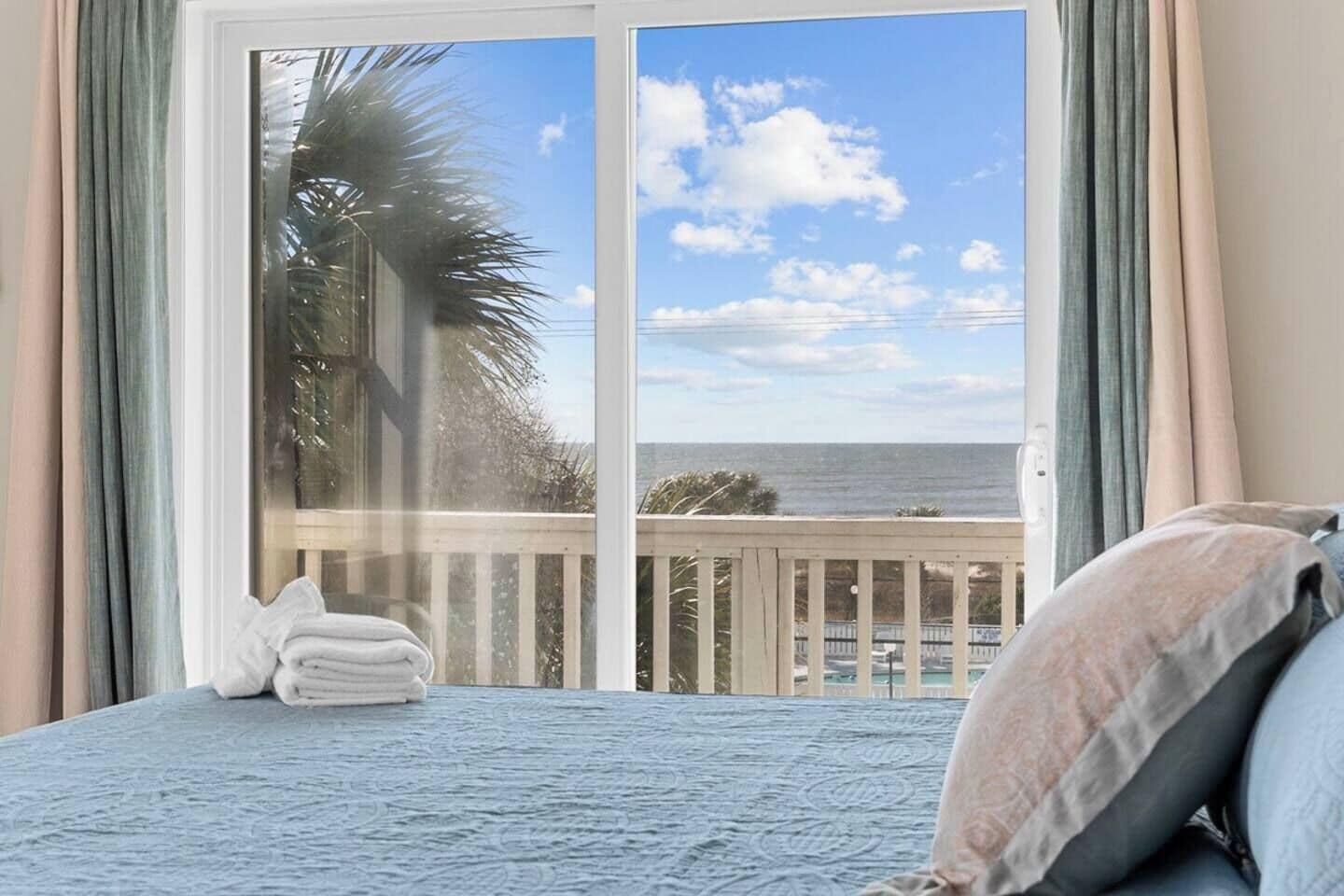 Une chambre avec vue sur l’océan, accessible par une porte-fenêtre coulissante, comprenant un lit avec des oreillers et une fenêtre avec des rideaux.