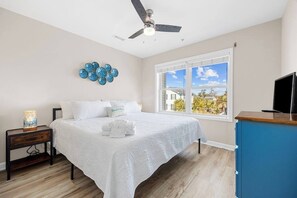 Un dormitorio con cama, mesita de noche, ventilador de techo, ventana con vistas y mueble para televisor.