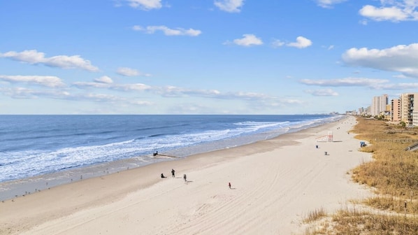 A wide sandy beach with ocean waves, a few people, and buildings in the background.