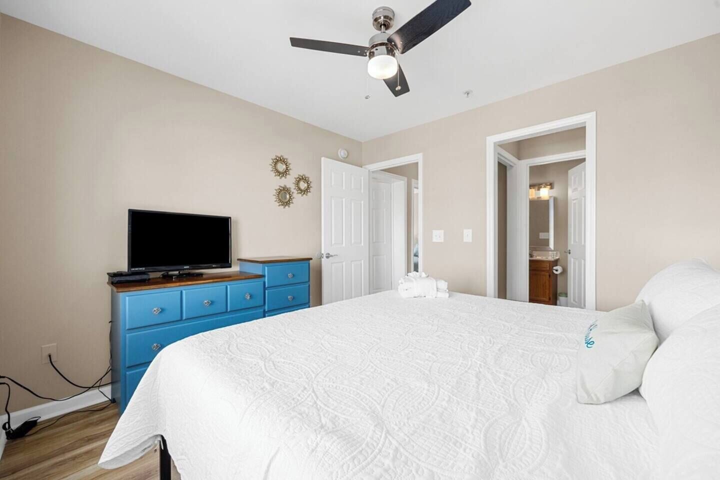 Een slaapkamer met een bed, een televisie op een dressoir, een plafondventilator en een deur naar een andere kamer.