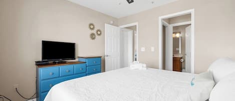 Quarto com cama, televisão sobre uma cômoda, ventoinha no teto e porta que dá para outro espaço.