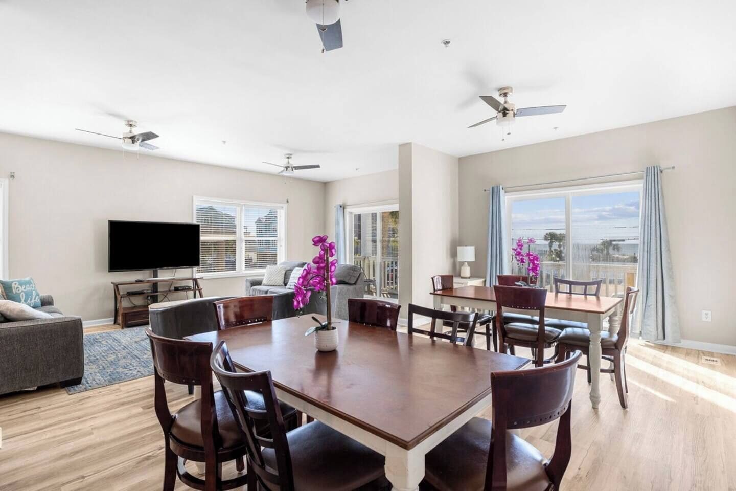 A spacious living room with a large dining table, comfortable seating, a flat-screen TV, and a balcony with a view.