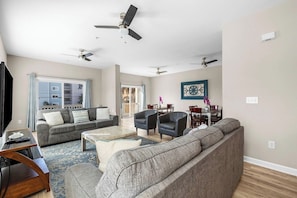 A spacious living room with a gray sofa, a coffee table, and a TV. There are ceiling fans, a dining area with chairs, and a large window with curtains.