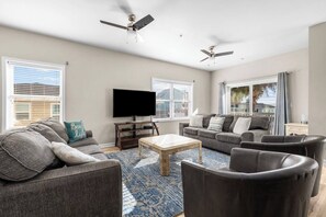A spacious living room with a flat-screen TV, two ceiling fans, and a large window providing a view of the outdoors.