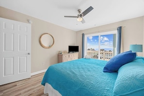 Un dormitorio con cama, ventilador de techo, televisión, cómoda, ventana con vistas y puerta.