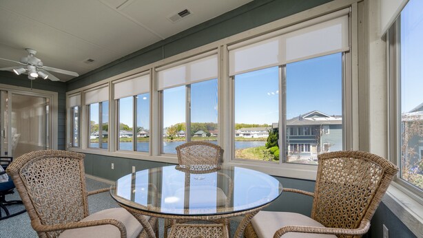 A sunroom with a glass table surrounded by wicker chairs, large windows, and a view of a lake and houses. - Condo with elevator and POND VIEWS! - 12 Pondview Lane (Lewes)