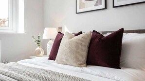 Un letto rifatto con copriletto bianco e cuscini, una lampada da comodino e un quadro appeso al muro.