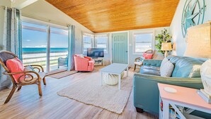 A spacious living room with a wooden ceiling, large windows, a television, and comfortable seating. - Primrose Cottage - S Nags Head MP 19, Oceanfront, Steps to the Water, Well Equipped, Pets OK (Nags Head)