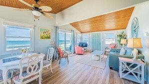 A spacious living room with a wooden ceiling, large windows offering a view of the ocean, a dining area with a white table and chairs, and a cozy seating area with a green sofa and a television. - Primrose Cottage - S Nags Head MP 19, Oceanfront, Steps to the Water, Well Equipped, Pets OK (Nags Head)