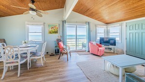 A spacious living area with a wooden ceiling, a dining table with chairs, a pink sofa, a television, and a view of the ocean. - Primrose Cottage - S Nags Head MP 19, Oceanfront, Steps to the Water, Well Equipped, Pets OK (Nags Head)