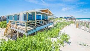 A beach house with a wooden deck and a covered porch, situated on stilts over the sand. - Primrose Cottage - S Nags Head MP 19, Oceanfront, Steps to the Water, Well Equipped, Pets OK (Nags Head)