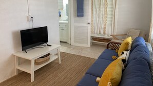 Ein Wohnzimmer mit Fernseher auf einem weißen Schrank, einem blauen Sofa mit gelben Kissen, einem Holzstuhl und einem weißen Konsolentisch.