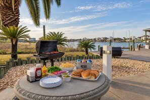 Ein Grillbereich im Freien mit Blick auf ein Gewässer, eine Holzterrasse und ein Steintisch mit Essen und Beilagen.