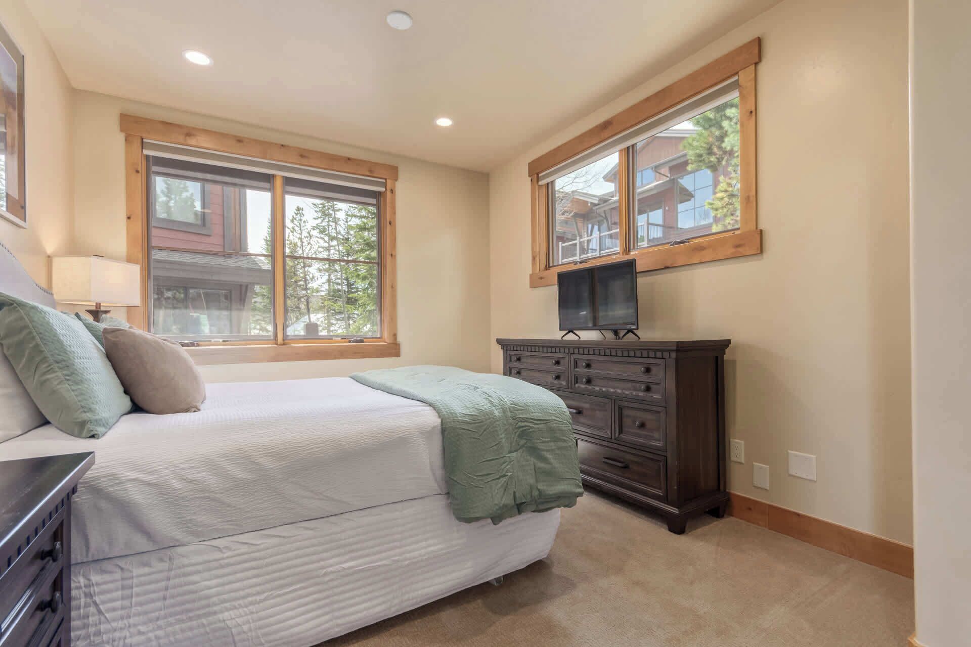 A bedroom with a bed, dresser, TV, and large windows.