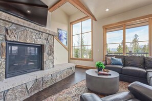 A living room with a stone fireplace, a large window with a view of trees and mountains, a leather sofa, and a round coffee table. - Incredible 4BR Tranquil Setting & Space to Spare! (Silverthorne)