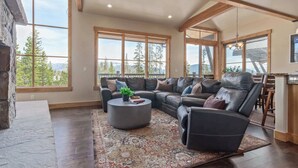Ein geräumiges Wohnzimmer mit einer großen Eckcouch, einem Couchtisch und Blick auf Bäume und Berge durch große Fenster.