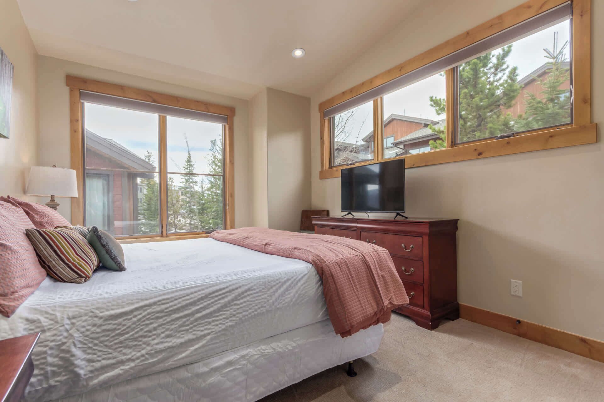 A bedroom with a bed, a TV on a dresser, and large windows with a view of trees and buildings.