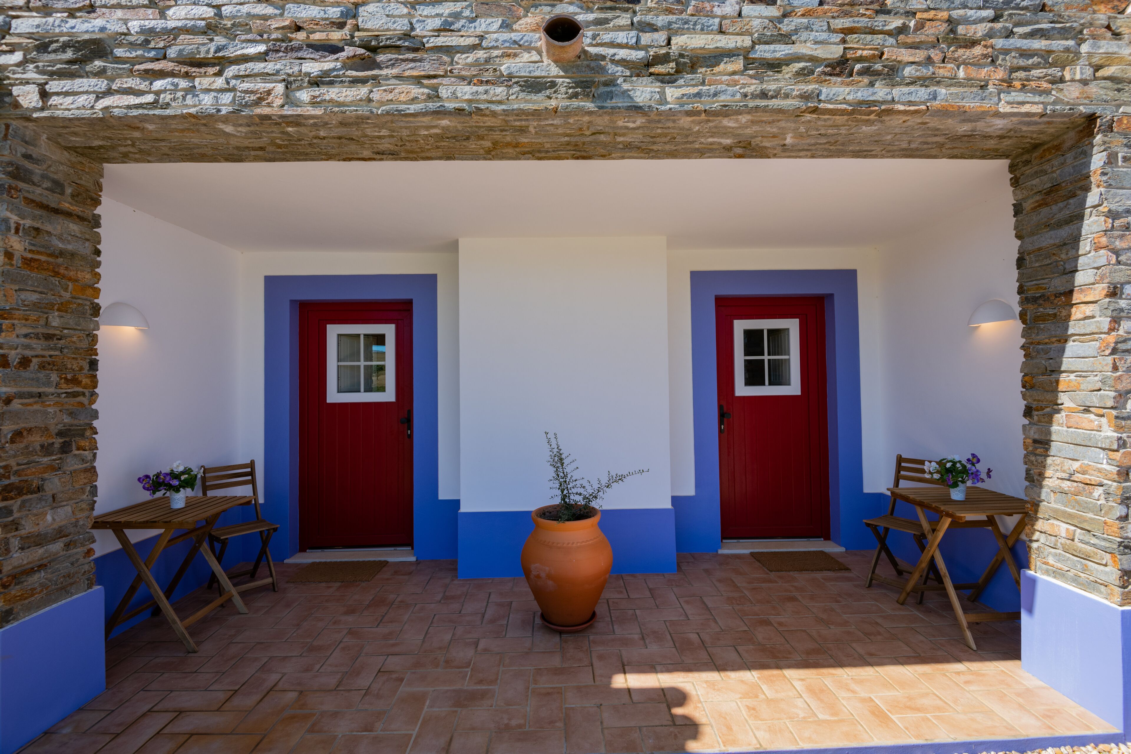 A covered outdoor area with two red doors, a large terracotta pot, and wooden furniture.