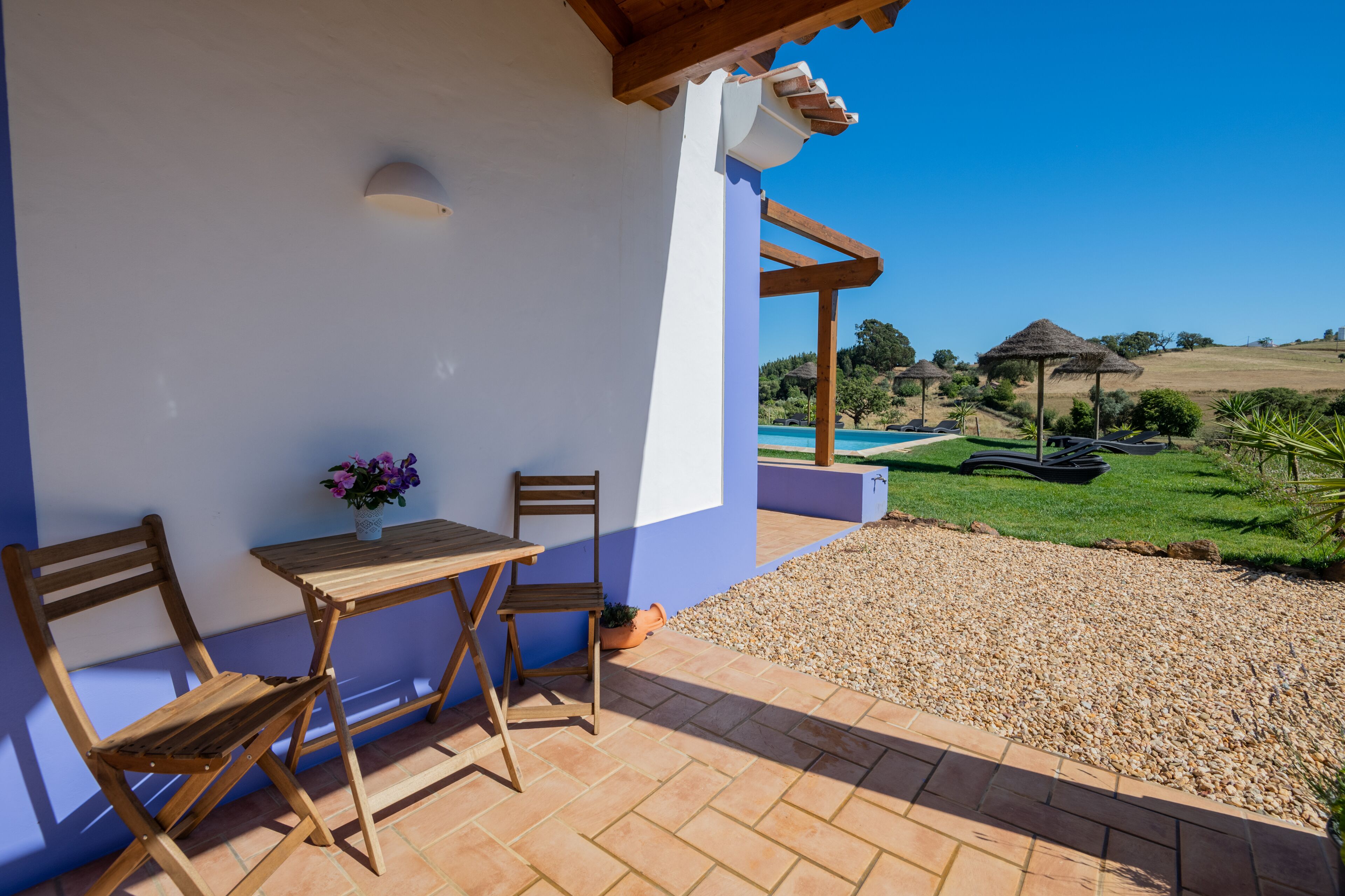 A patio with a table and chairs, a potted plant, and a view of a swimming pool and landscape.
