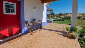 Eine überdachte Terrasse mit einer roten Tür, einem Holztisch und -stühlen sowie Blumentöpfen.