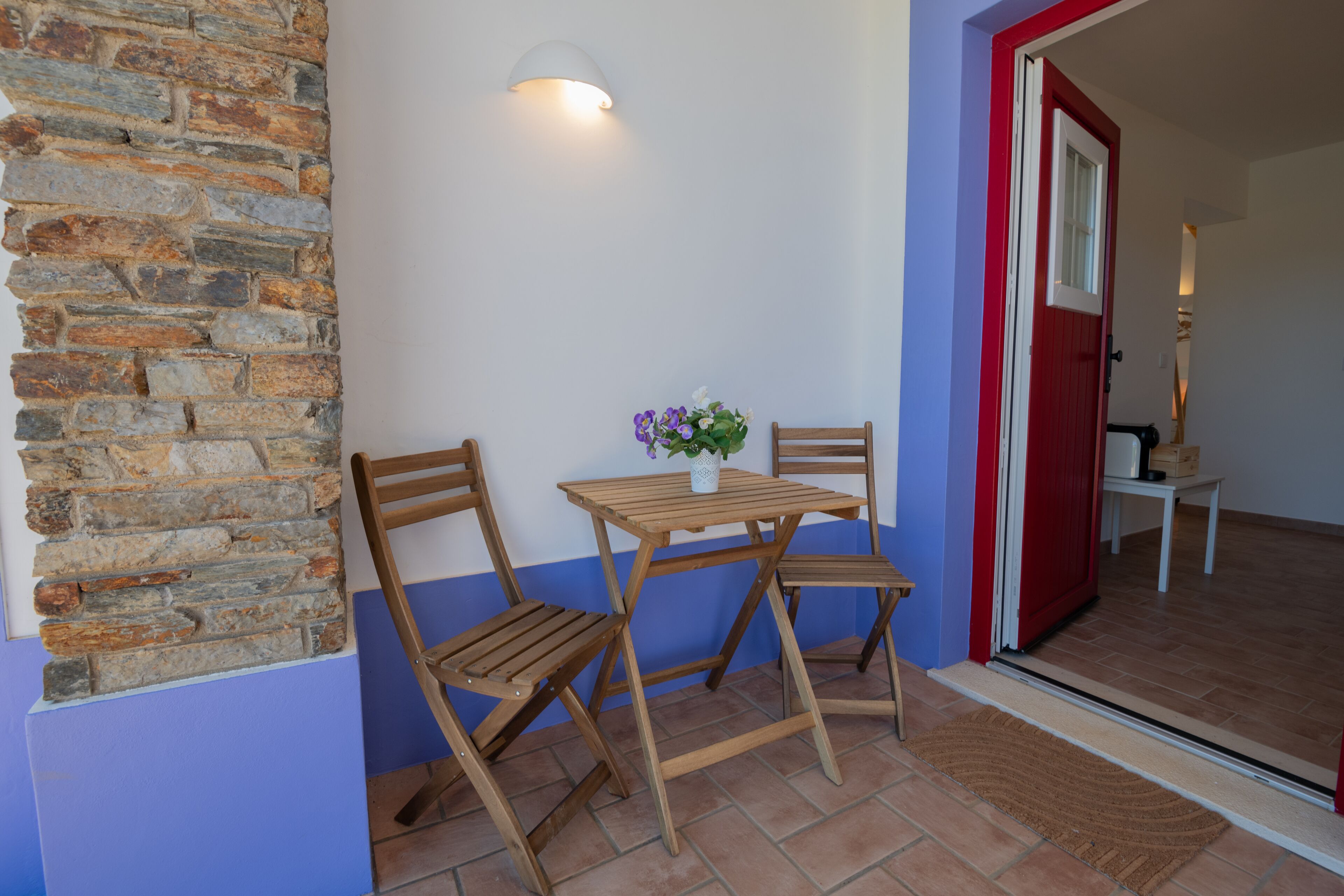 Una habitación con pared de piedra, sillas de madera, una mesita y una puerta con marco rojo.