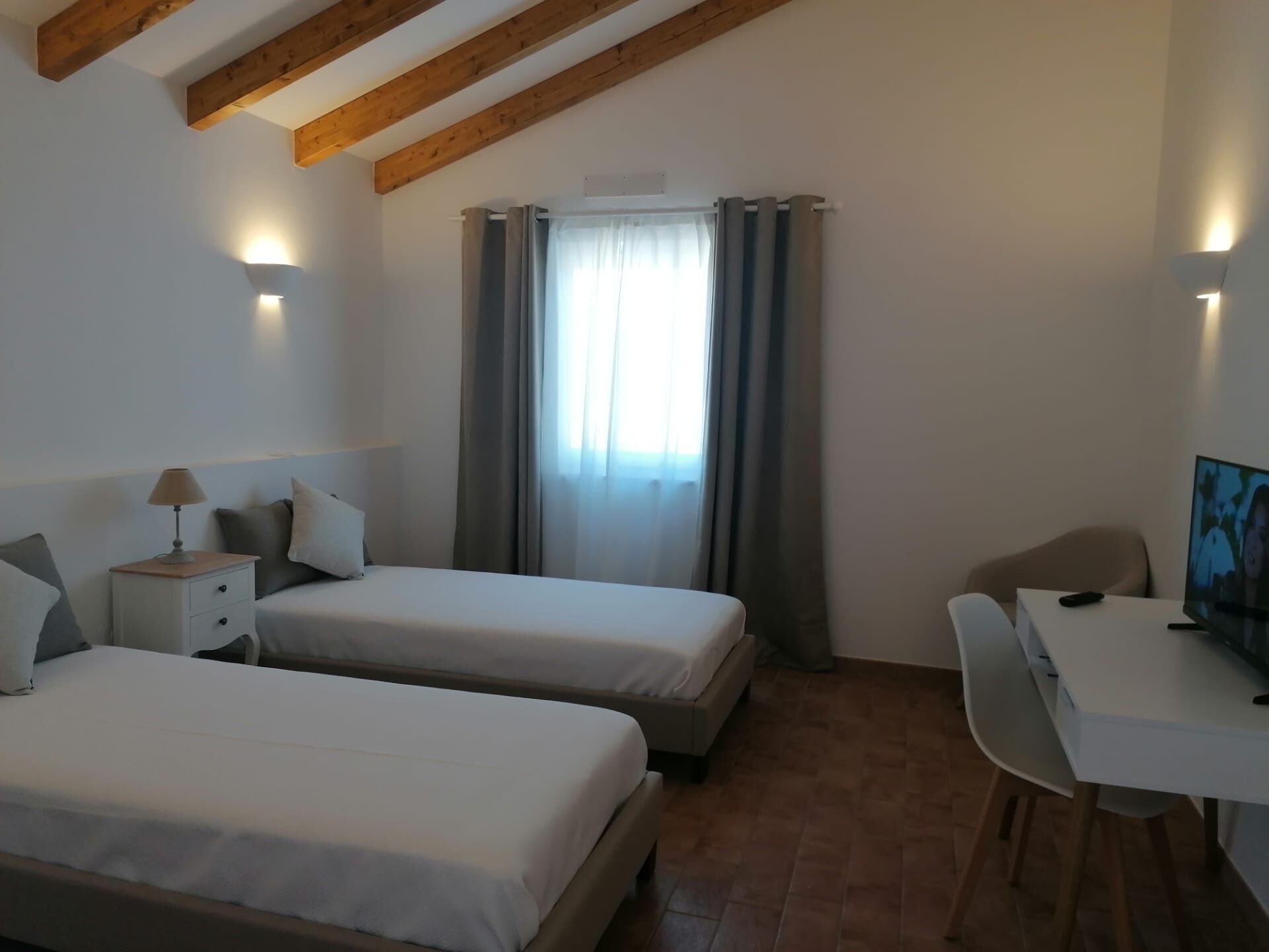 A bedroom with two beds, a desk, and a chair. There is a window with curtains, a lamp on the bedside table, and a TV on the desk.