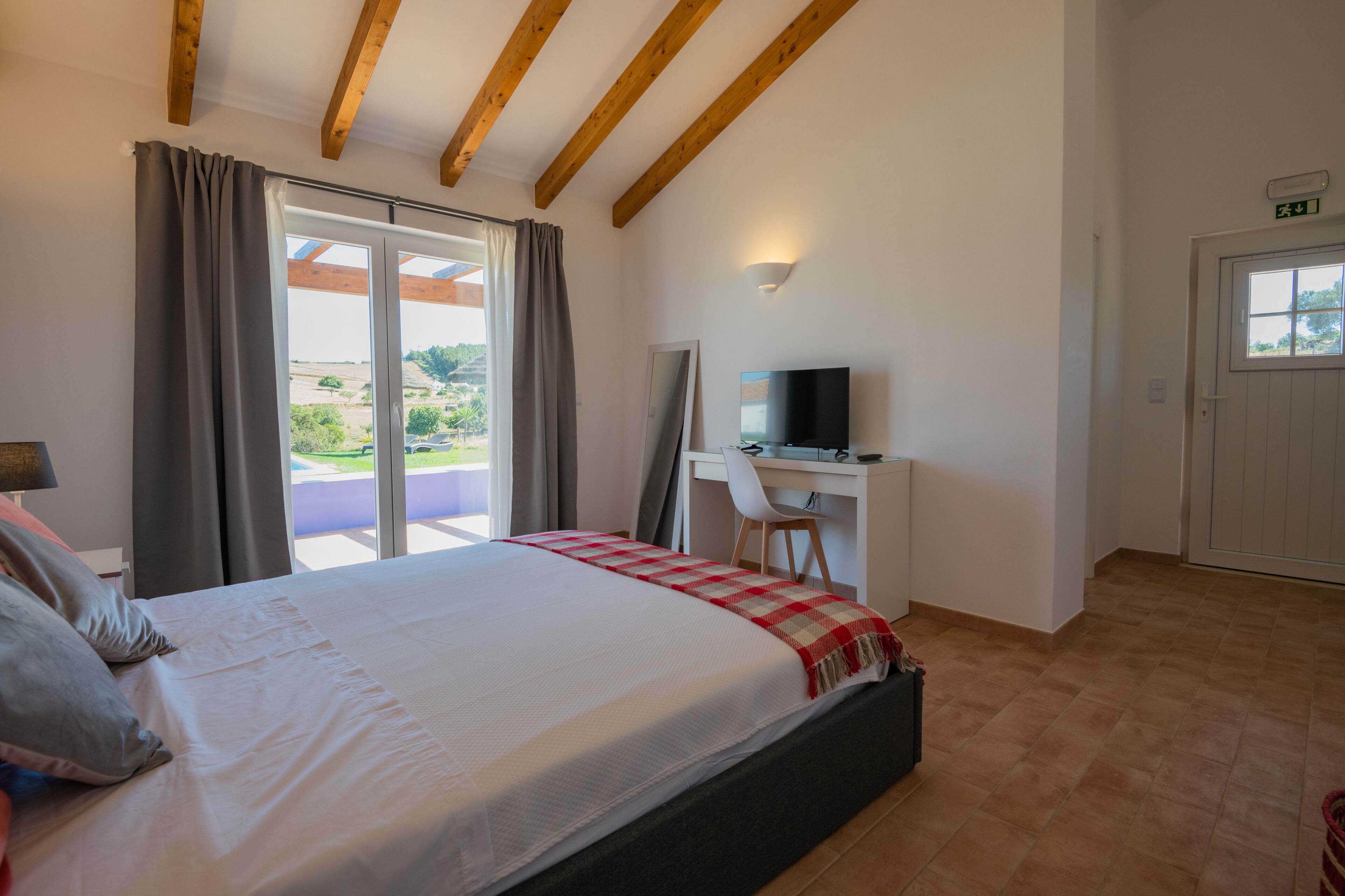 A bedroom with a bed, a desk, a chair, a TV, and a sliding glass door leading to a balcony.