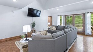 Un salon spacieux avec un grand canapé d’angle gris, un téléviseur à écran plat fixé au mur et de grandes fenêtres dotées de rideaux.