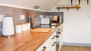 A modern kitchen with wooden countertops, a built-in toaster, and a microwave. There are shelves above the counter holding cups and a lamp. - Gerry's Place (Church Stretton)